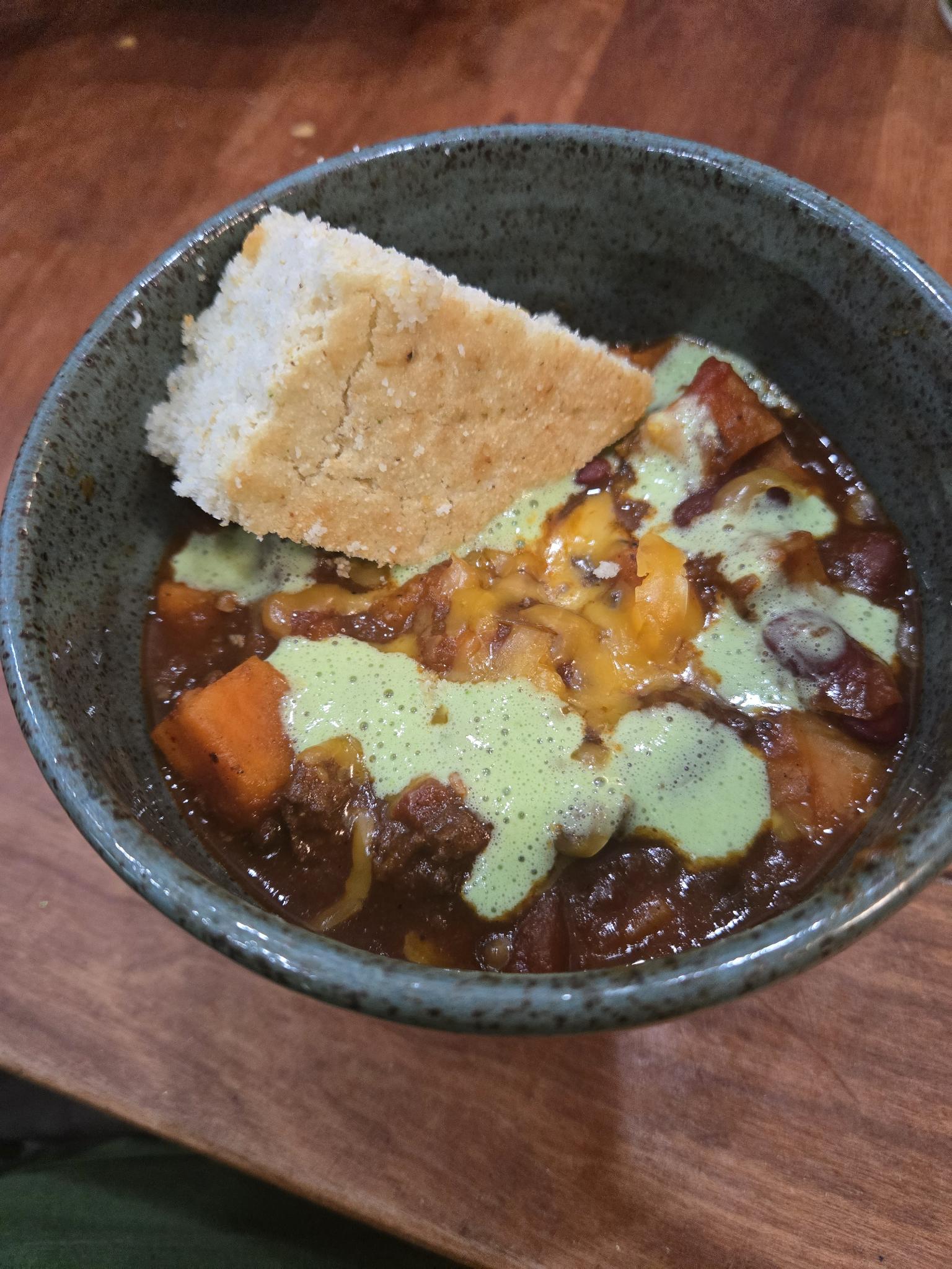 A green bowl full of beef and sweet potato chili. Topped with shredded cheese, chili lime cilantro yogurt sauce, and cornbread.